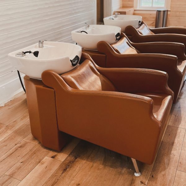 Three brown salon chairs with white washbasins lined up in a bright room with wooden floors and walls.