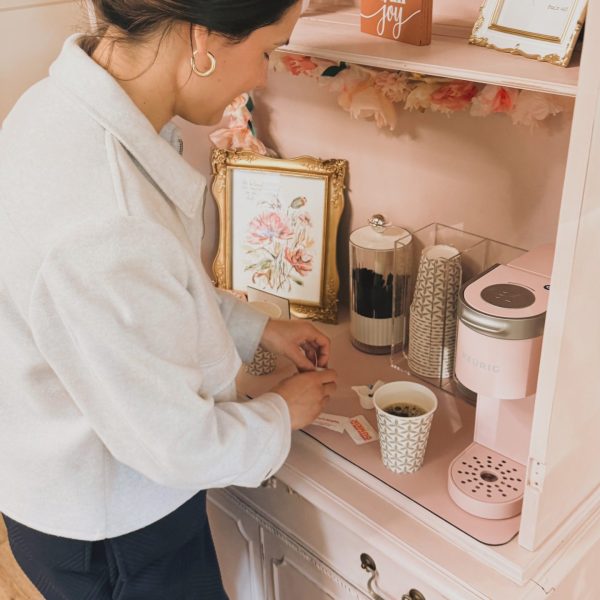 Woman prepares coffee at a pastel coffee station with art, mugs, and a “count it all joy” sign.