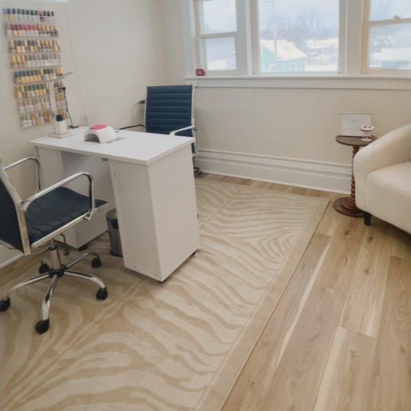 Modern nail salon room with a white desk, two blue chairs, a display of nail polish, and large windows.
