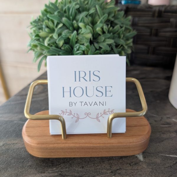 A small "IRIS HOUSE BY TAVANI" sign in a wooden and brass holder, with a potted plant behind it.